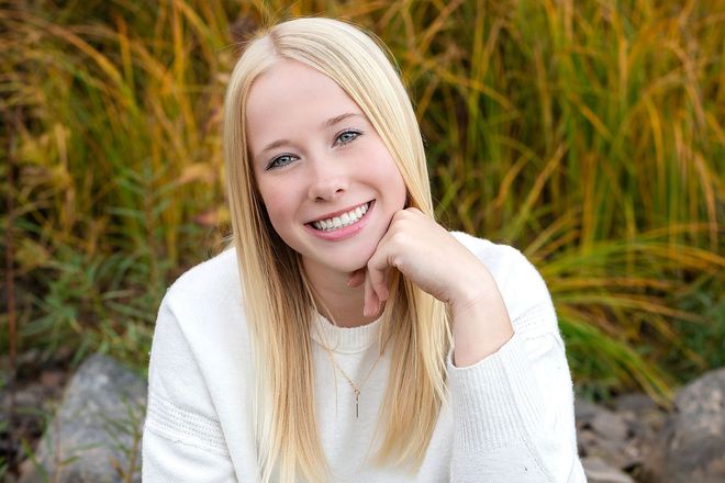 Smiling blonde woman in a white sweater posing outdoors among tall grass and rocks