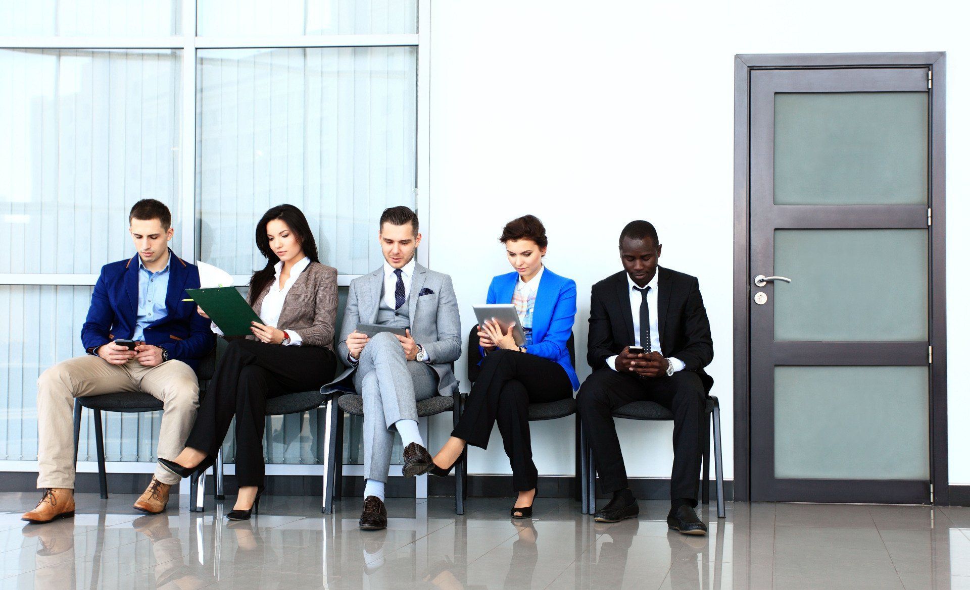 Five people sit on a bench in a bright waiting area, looking down at their phones or papers.