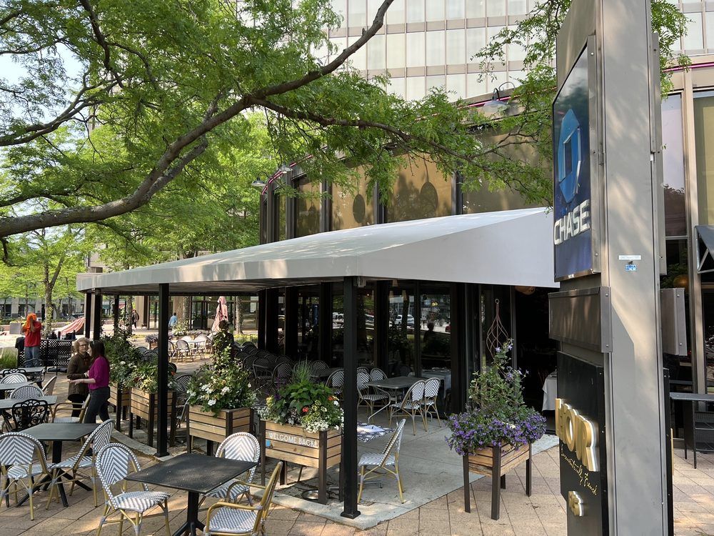 Restaurant patio with white awning, tables, chairs, and potted plants. Building and trees visible.