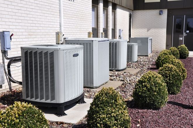 Air conditioning units lined up outside a brick building, with bushes and gravel in front.