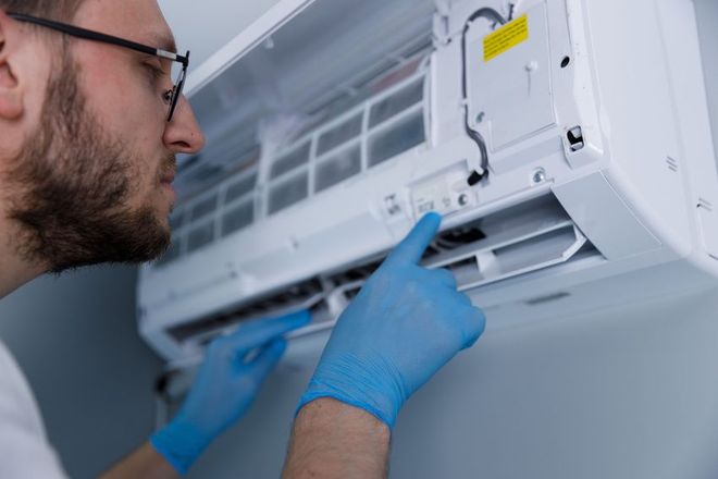 A person with blue gloves examines an open white air conditioner unit mounted on a wall.