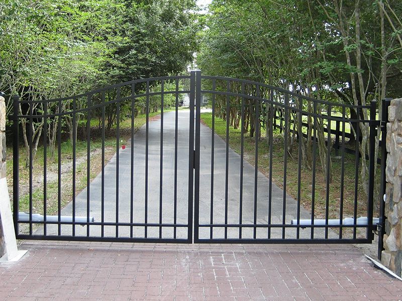 A black gate is open to a driveway surrounded by trees