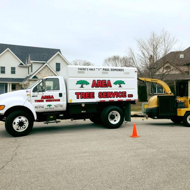 A white Area Tree Service dump truck towing a wood chipper parked on a residential street in front of two houses.