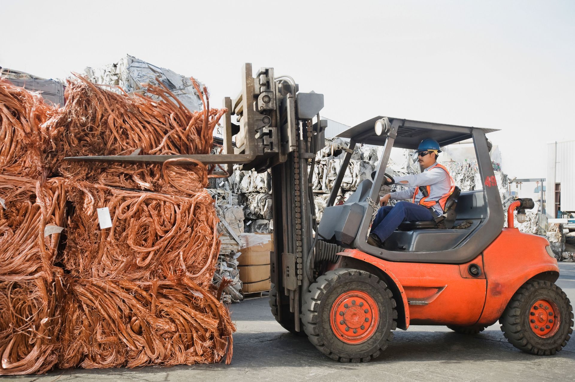 Forklift operator moving a bale of copper wire at a scrap yard. The forklift is orange.