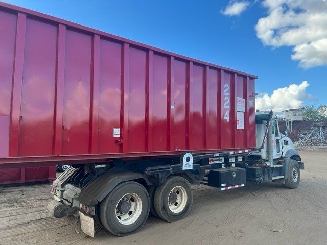 Red dumpster on a truck, parked on a gravel surface under a cloudy sky.