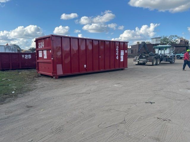Red dumpster being loaded by truck on a paved lot with a cloudy sky.
