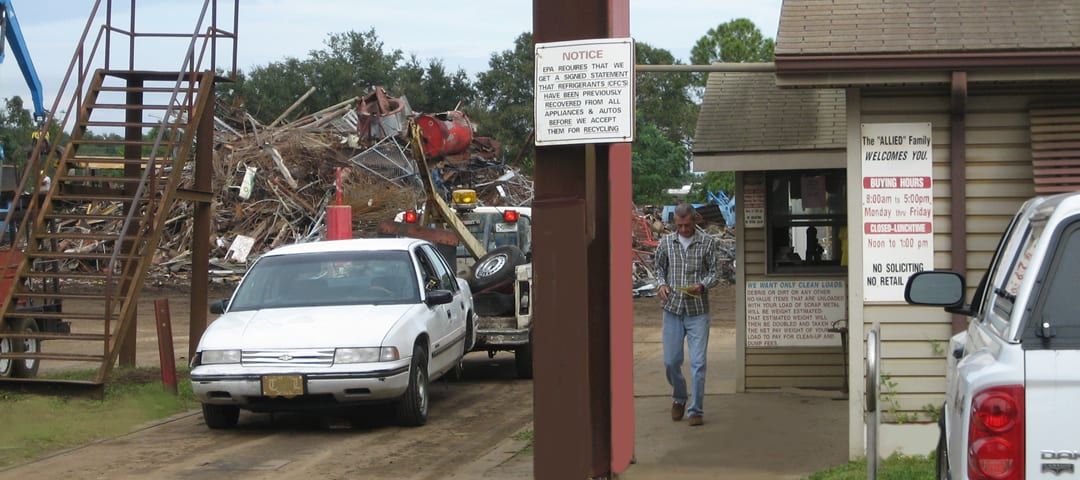 A junkyard with a white car entering, a man walking, and piles of scrap metal in the background.