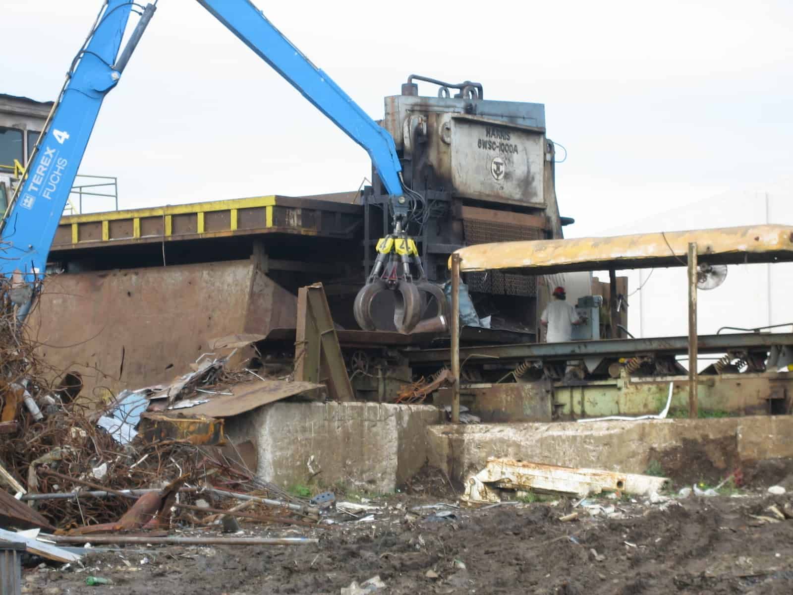 A blue crane with a claw-like grabber is lifting scrap metal into a recycling machine.