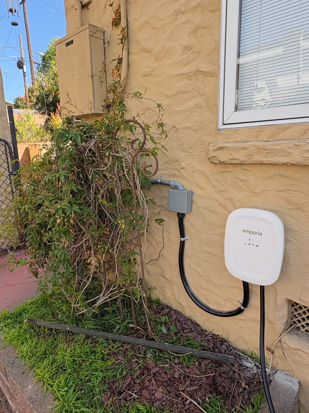 A white electric vehicle charger is mounted on a textured tan building next to overgrown greenery.