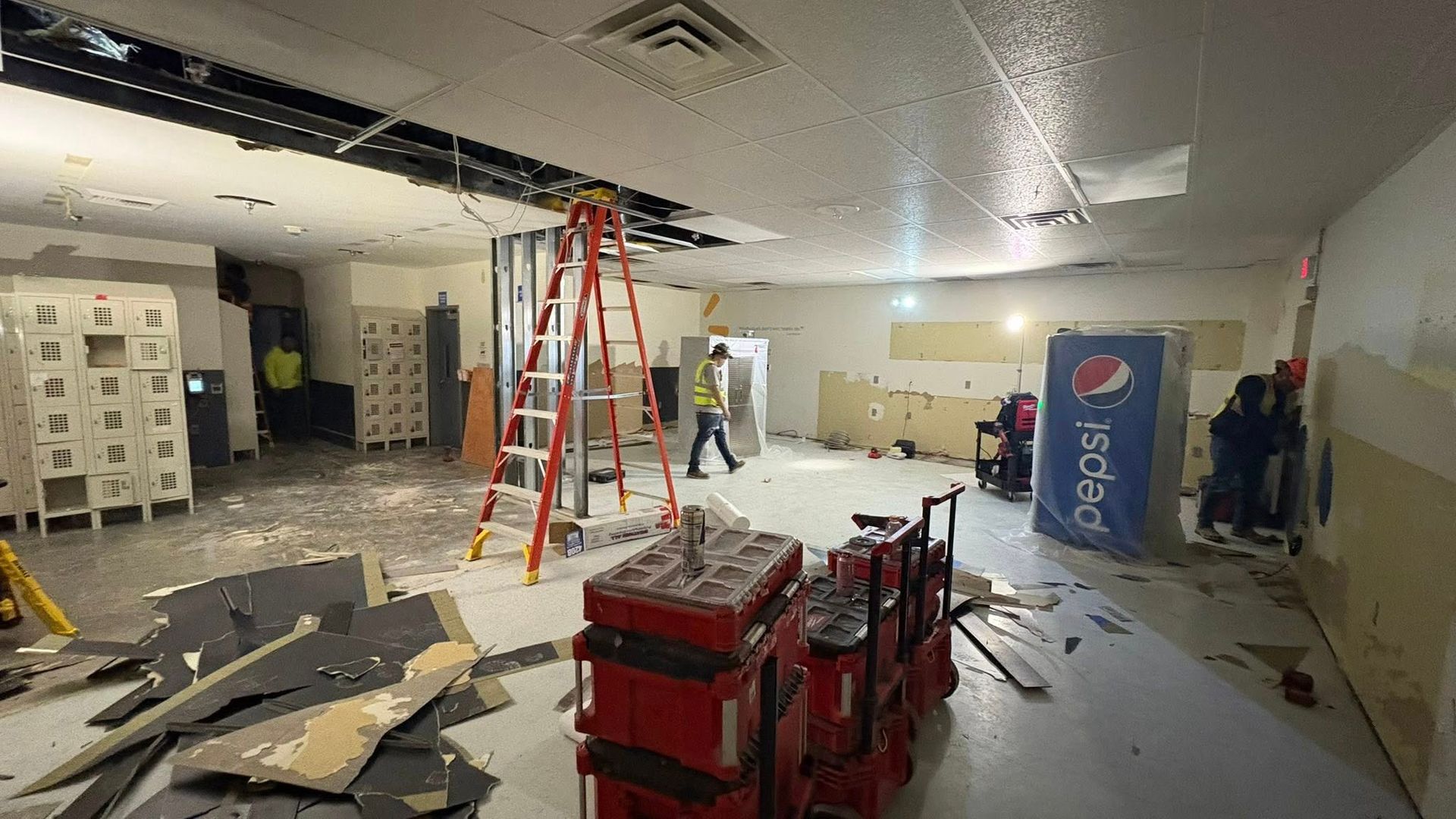 Construction site: Room being renovated. Workers, ladder, debris, Pepsi machine. White, gray, red, and yellow.