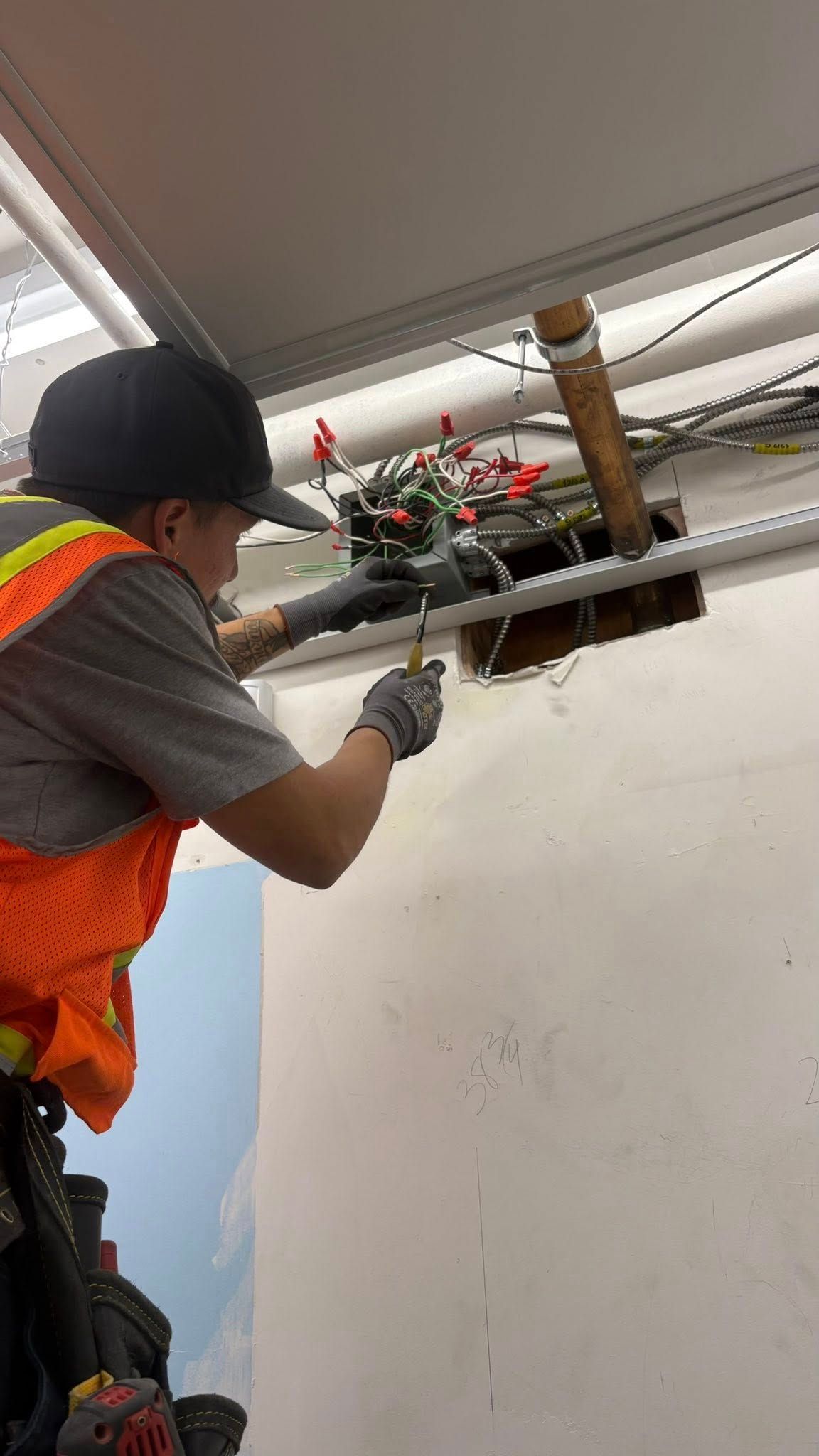 Person in safety vest and cap, working on exposed wiring in ceiling.