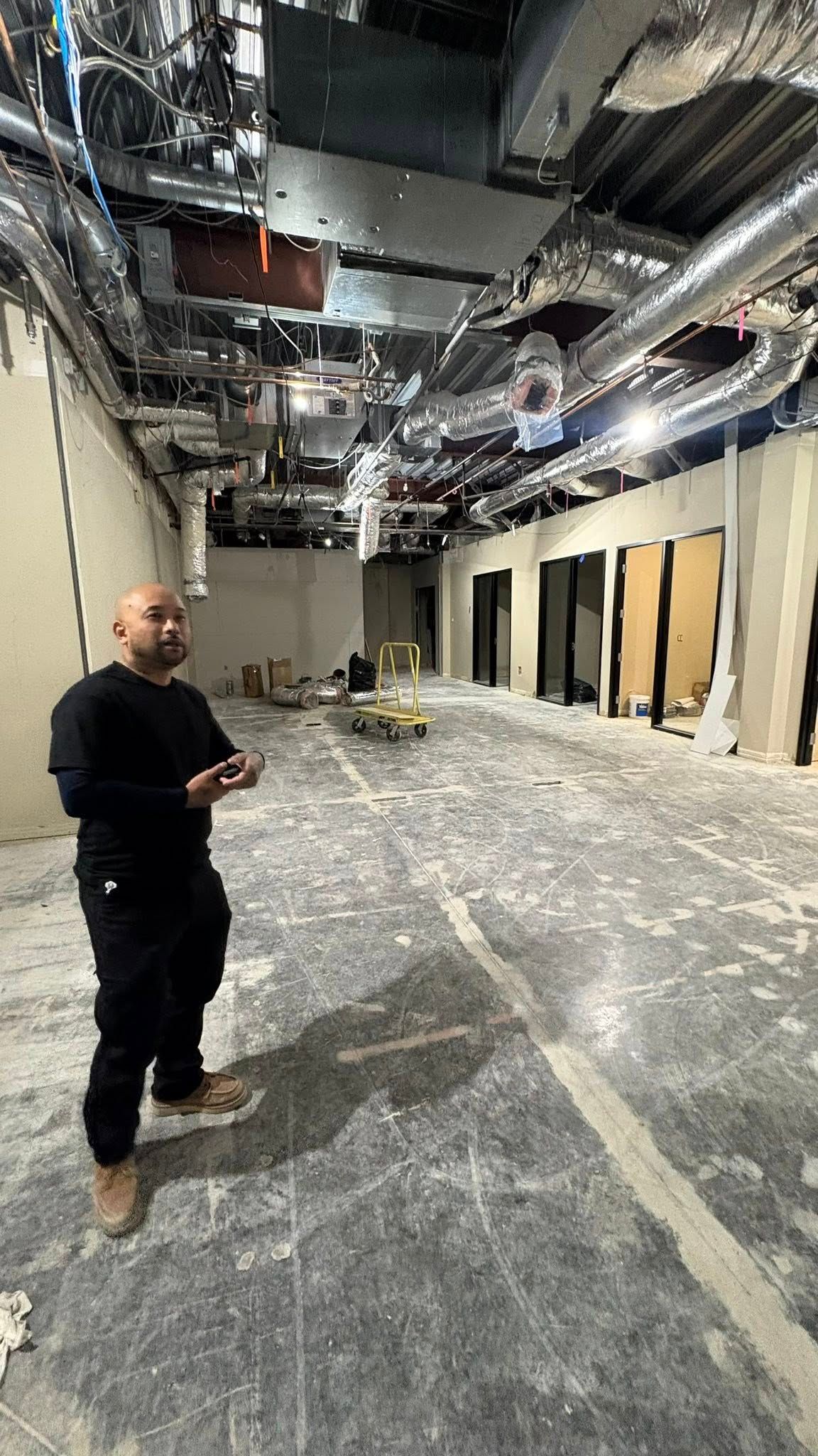 Man in black clothing inspects a construction site with exposed ducts, unfinished walls, and concrete floor.