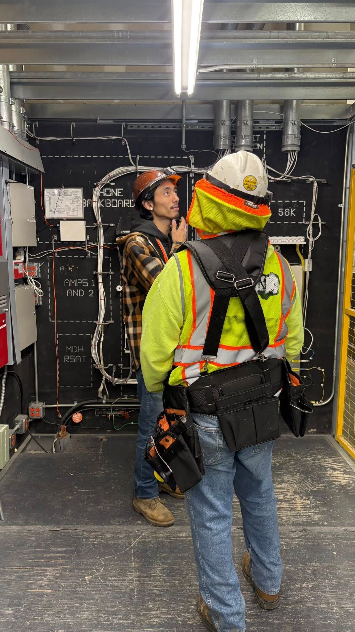 Two construction workers in a room with electrical wiring, one talking to the other.