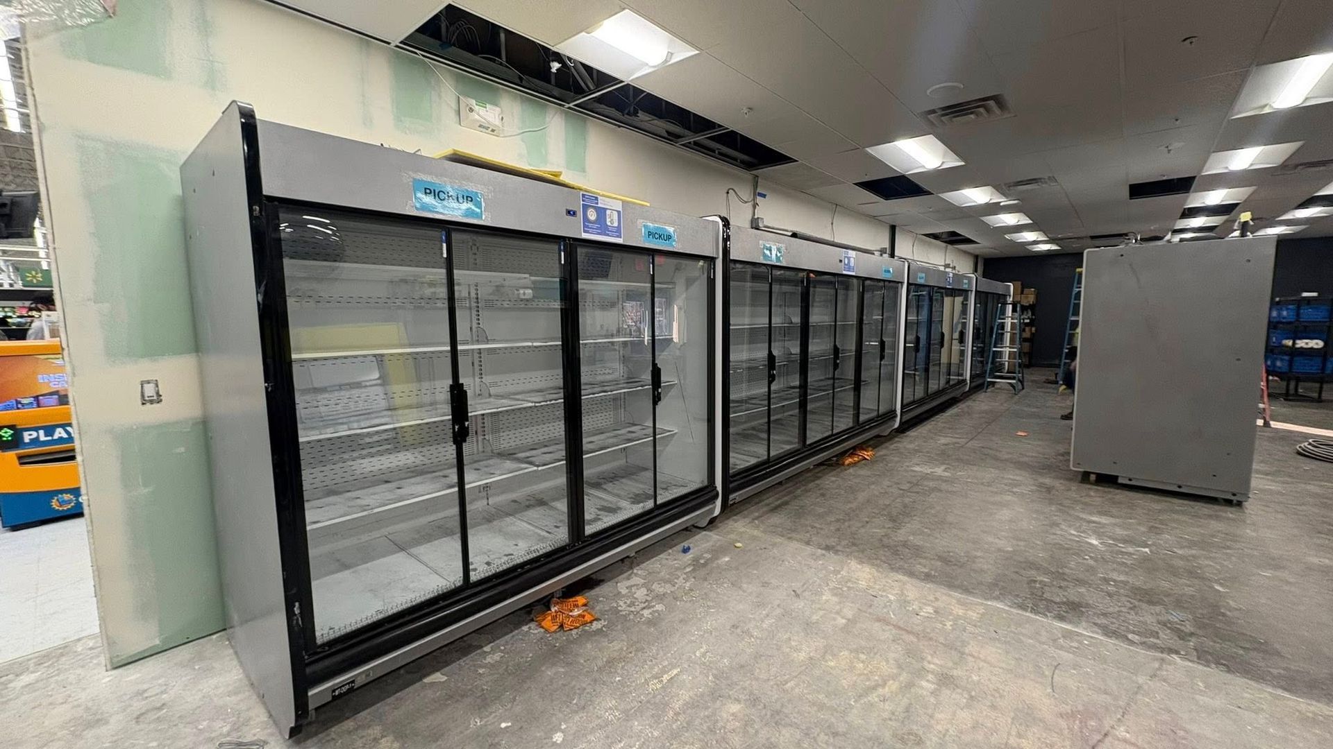 Row of empty commercial refrigerators in a store, with glass doors and stainless steel exterior. Interior undergoing renovation.