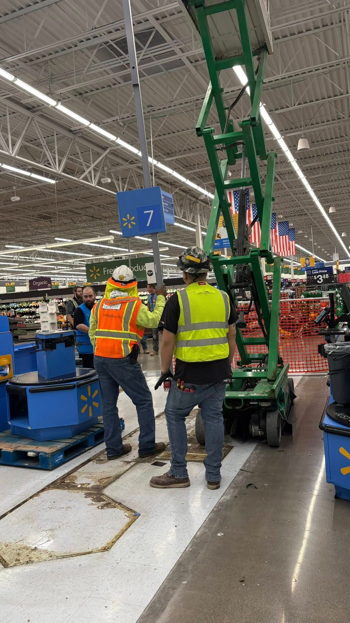 Two workers in a Walmart store near a lift, one with an orange vest and hard hat, the other in yellow.