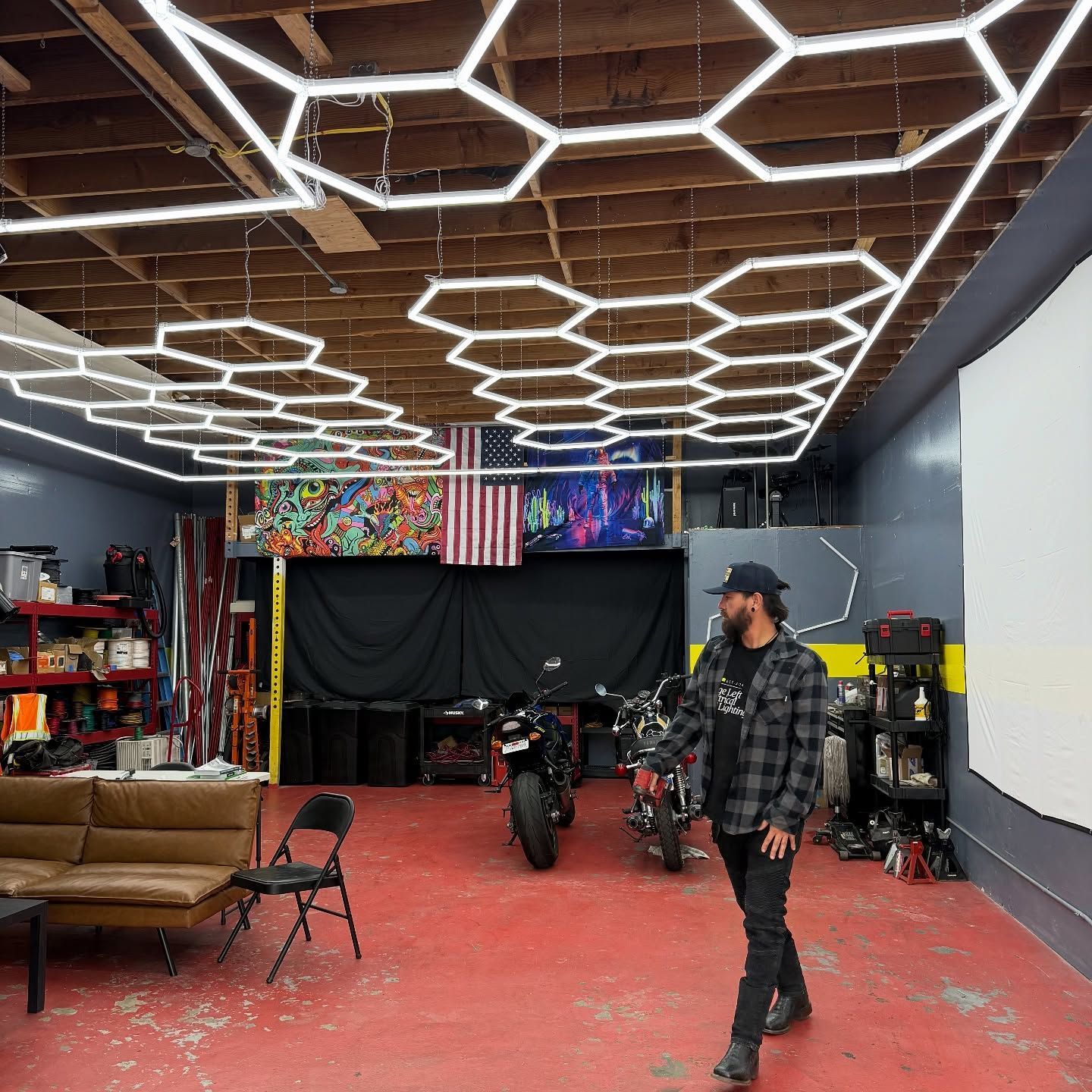 Man walking in a garage with hexagon-shaped lights on the ceiling, two motorcycles, and an American flag.