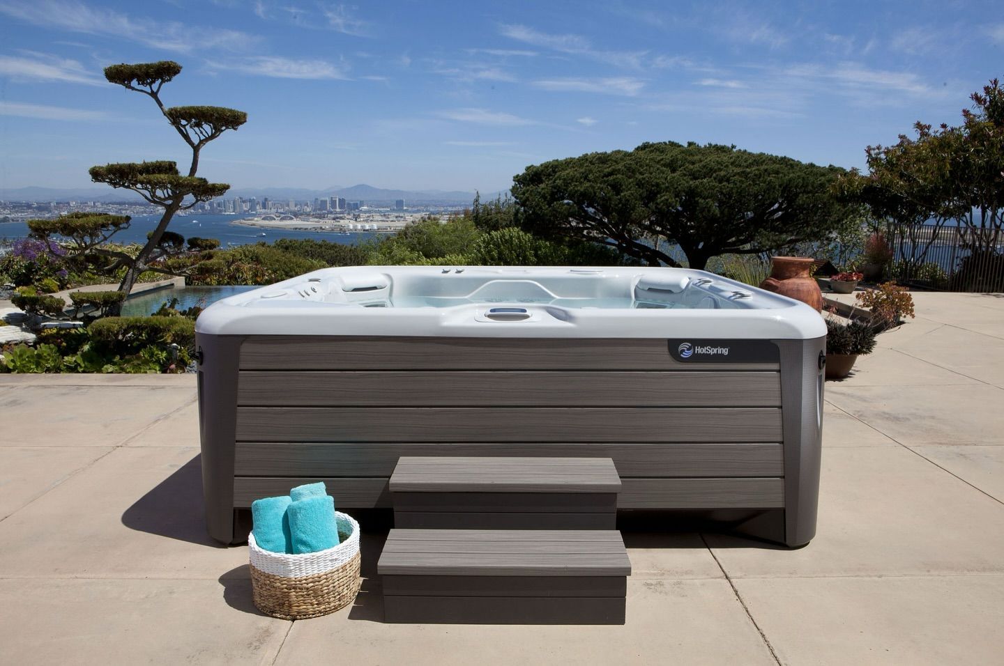 Hot tub on a patio with a view of a city skyline and water. Steps and a basket with towels are in front.