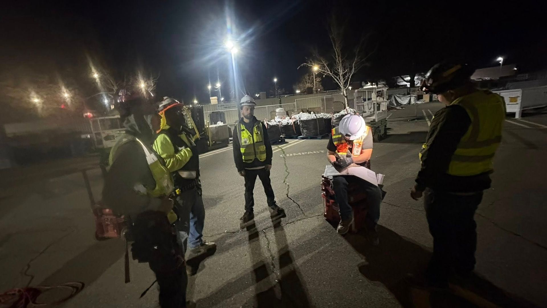 A group of people wearing safety vests and helmets stand on a dark road under bright lights at night.