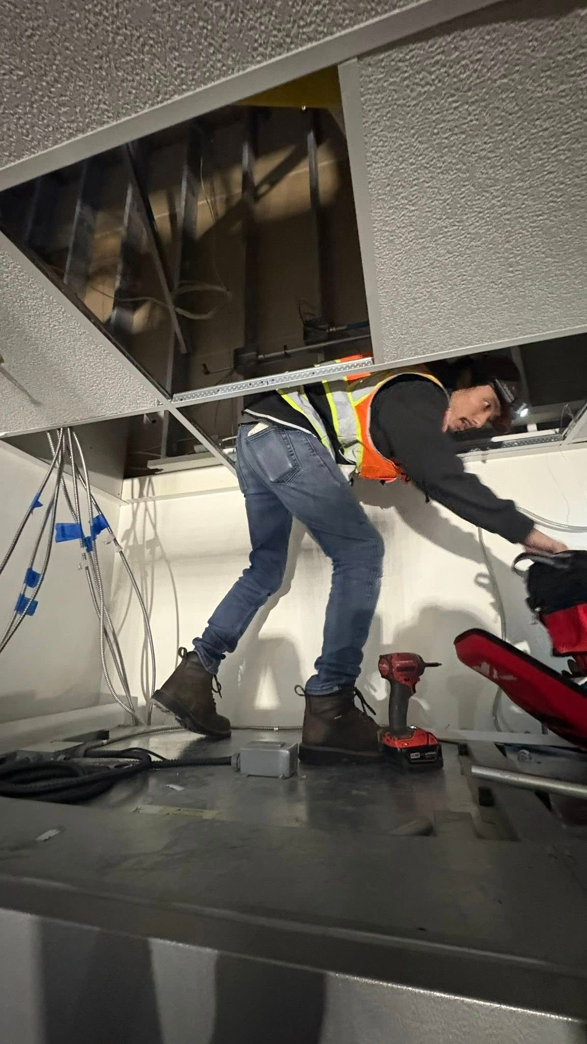 A worker in safety vest works on wiring above a ceiling. He is bent over, holding a tool, indoors.