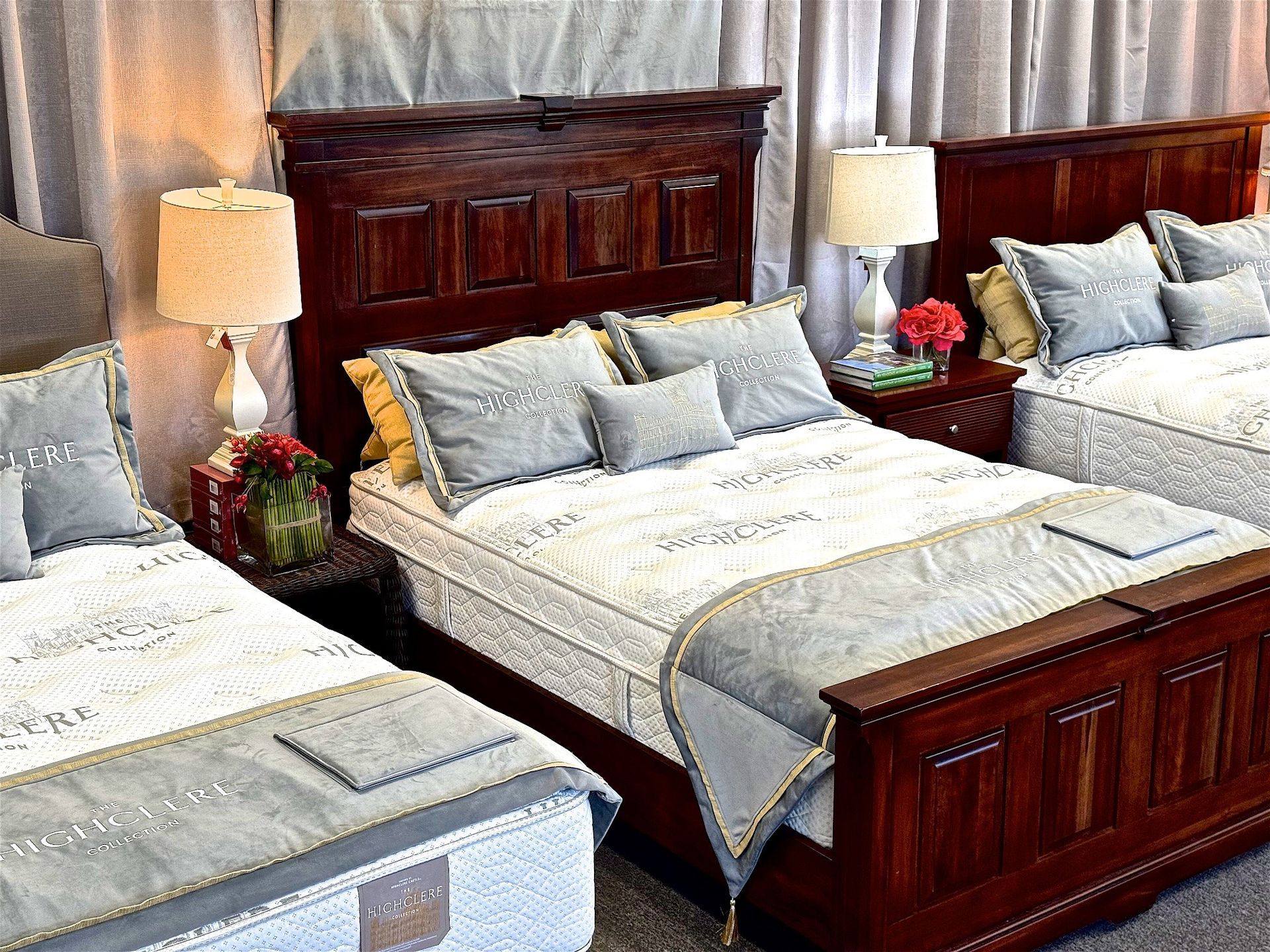 Three wooden beds on display, each with white mattresses, grey bedding, and matching lamps on nightstands.
