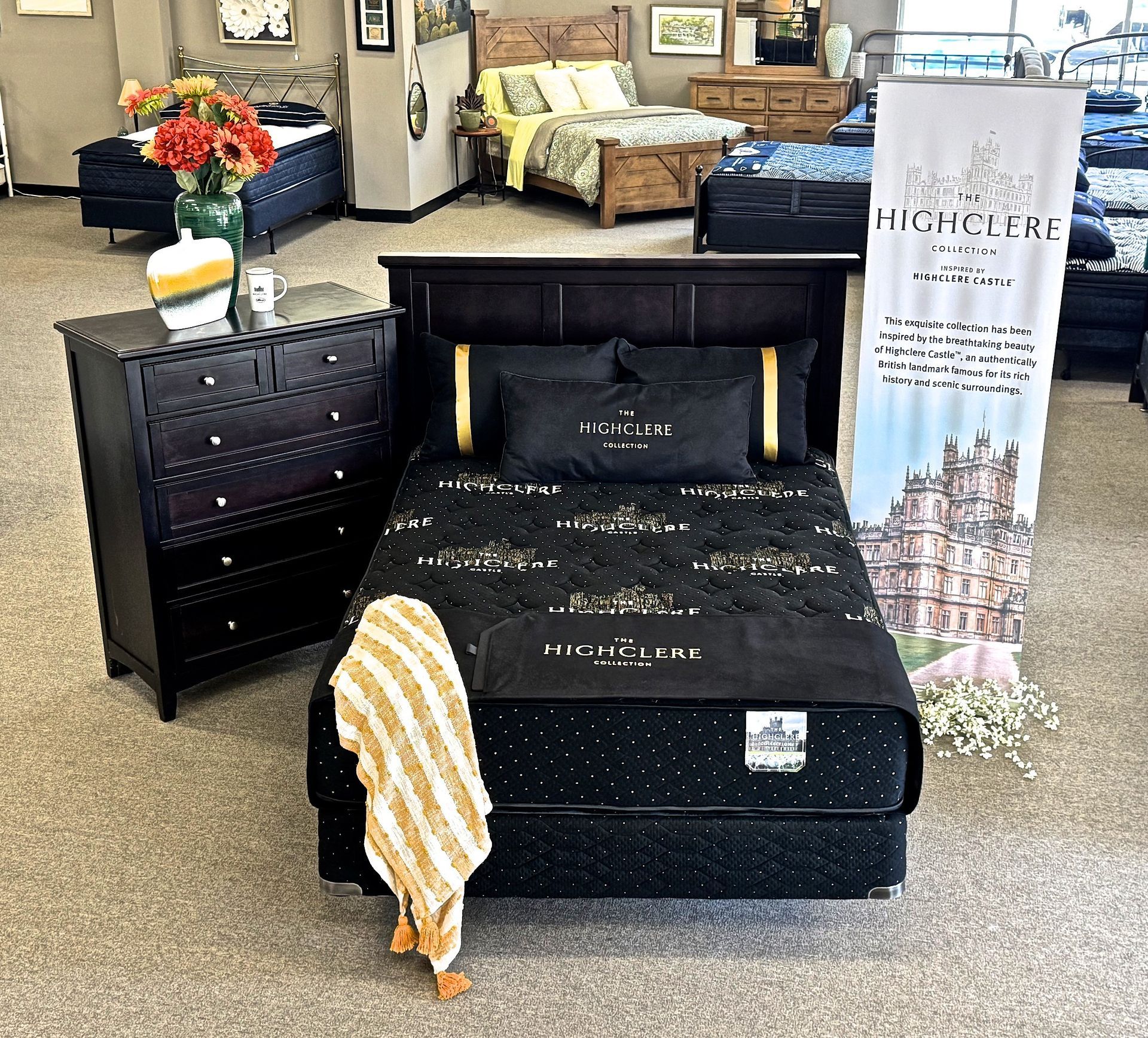 Bedroom furniture showroom display: black bed with bedding, dresser, banner, and other furniture.