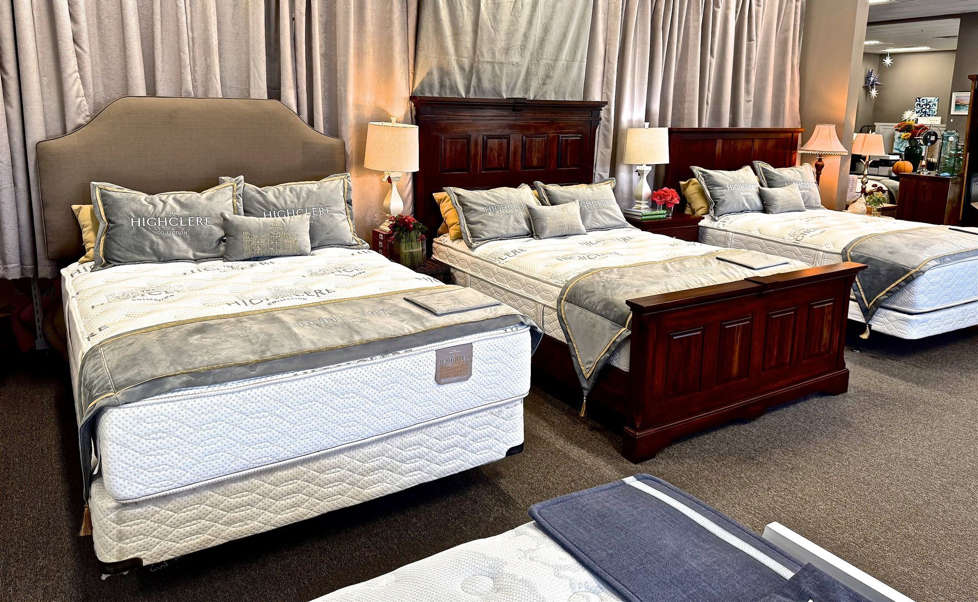 Three beds on display in a furniture store, each with linens and lamps, against draped curtains.