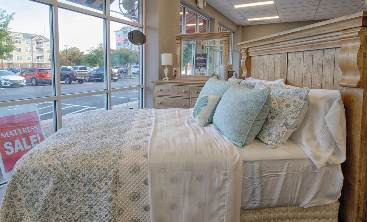 Bedroom furniture display in a store, bed with blue pillows and quilt, dresser, window view of cars.