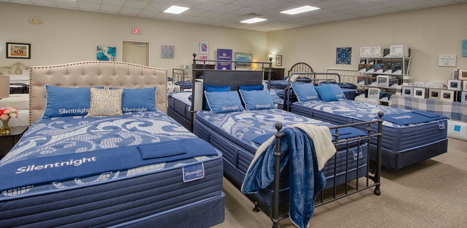 Mattresses displayed in a showroom. Beds are set up and dressed with blue linens. The room is well-lit.
