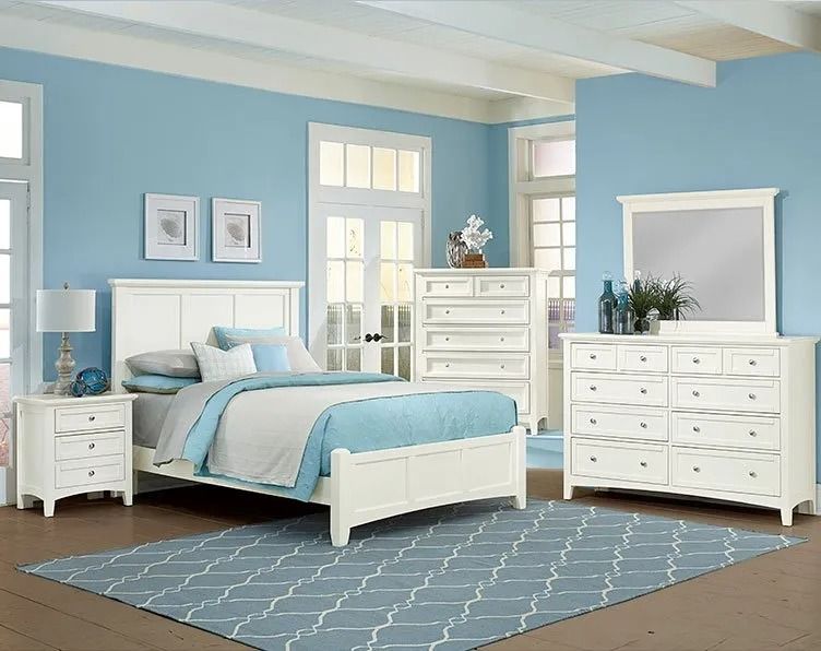 Bedroom furniture set against a blue wall; white bed, dresser, chest, and nightstand, blue rug.