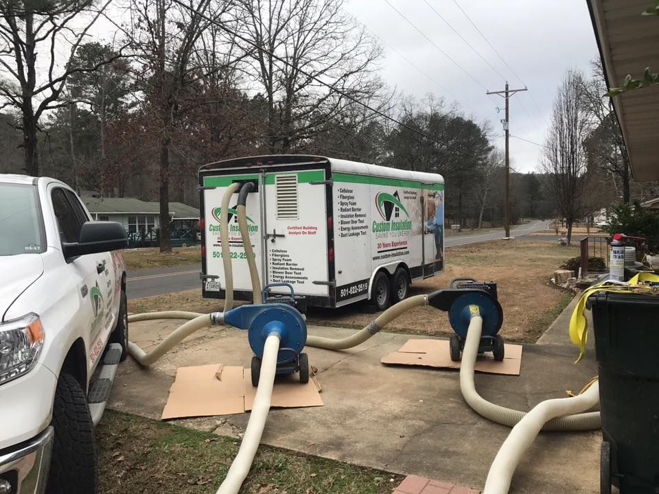 A white truck and trailer set up for insulation installation with two machines connected by hoses, on a residential driveway.
