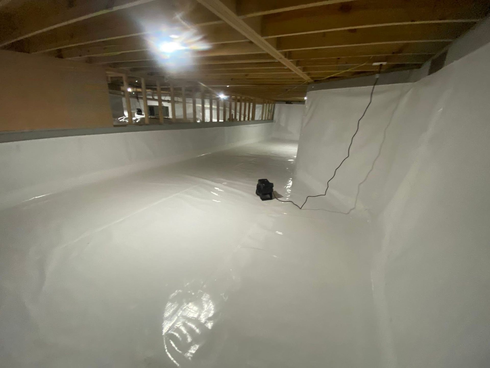 White-walled crawlspace with plastic sheeting, under wooden joists. A pump and electrical cord are visible.