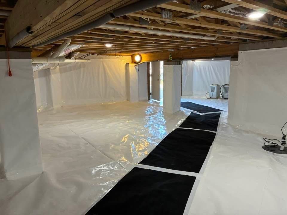 A crawl space with white vapor barriers covering walls and floor, featuring a black drainage mat path and a sump pump.