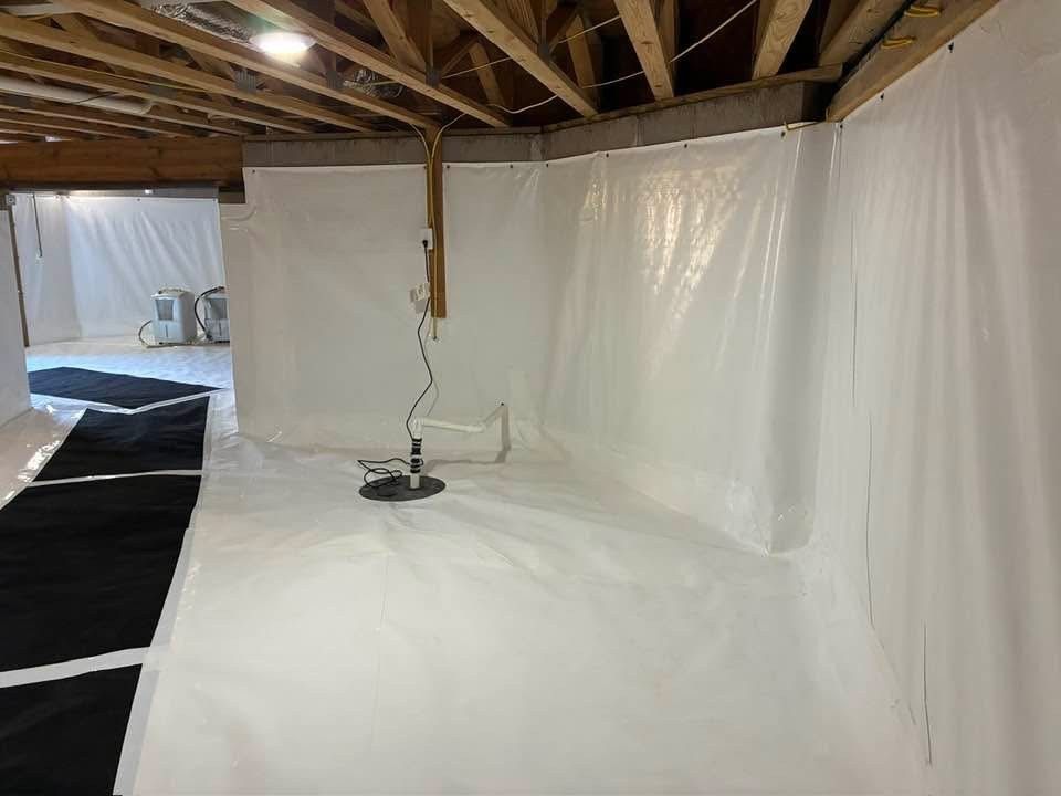 Crawl space lined with white vapor barrier, featuring a sump pump and overhead wooden floor joists.