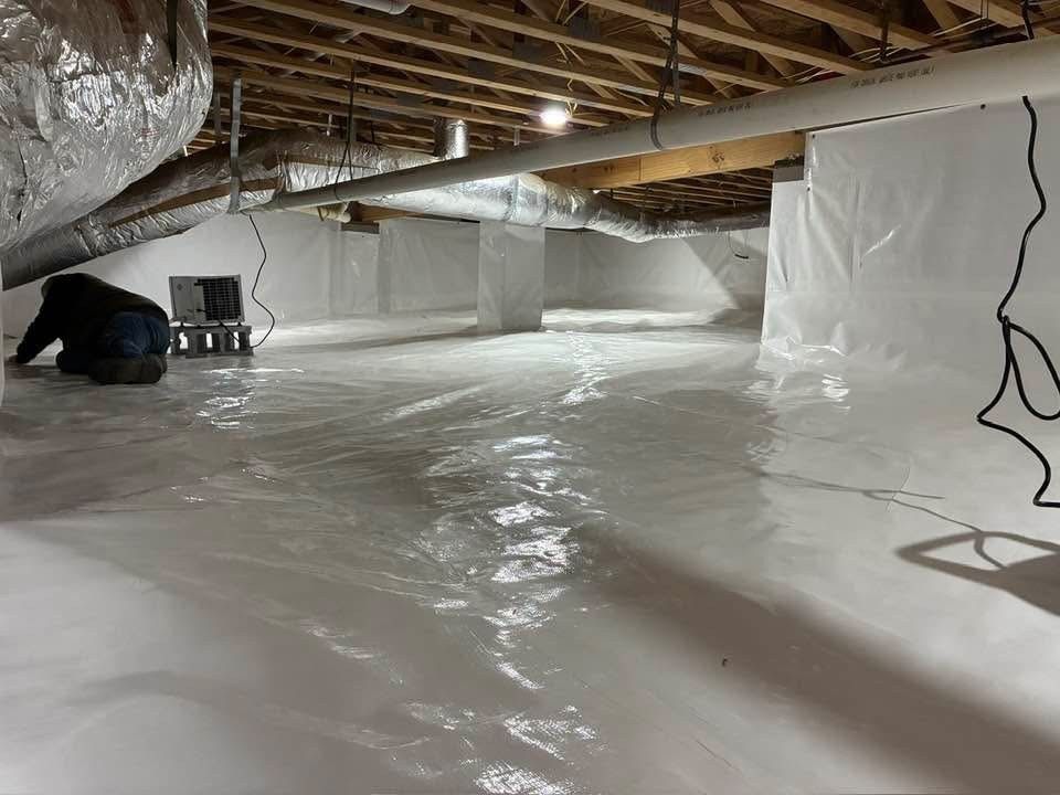 A person works in a crawl space encapsulated with white vapor barrier plastic and exposed wooden floor joists above.