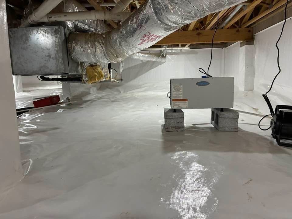 A crawlspace featuring a white vapor barrier on the ground, a silver dehumidifier on concrete blocks, and HVAC ductwork.