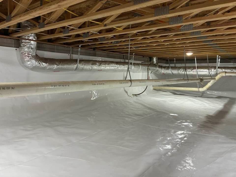 A clean crawl space with white vapor barrier flooring and walls, wooden floor joists, and suspended HVAC ducts and pipes.