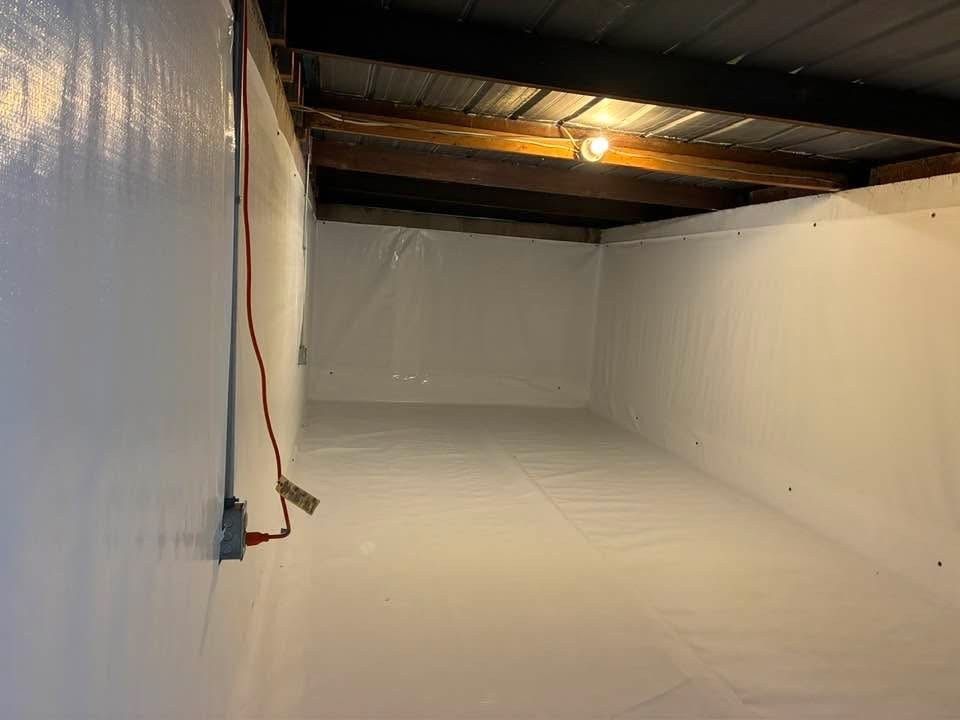 A crawl space interior lined with white vapor barrier plastic on the floor and walls under exposed wooden ceiling beams.