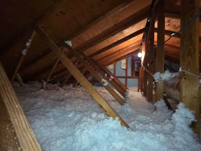 Attic with exposed wooden beams and rafters, insulated with white blown-in material around ductwork and support posts.