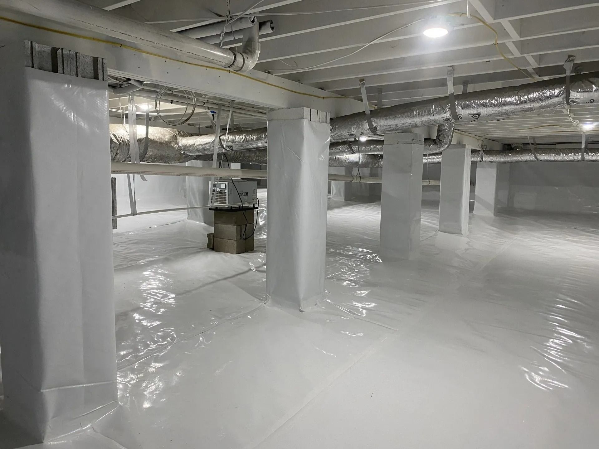 A crawl space encapsulated in white vapor barrier. Support columns, pipes, and ductwork are visible.