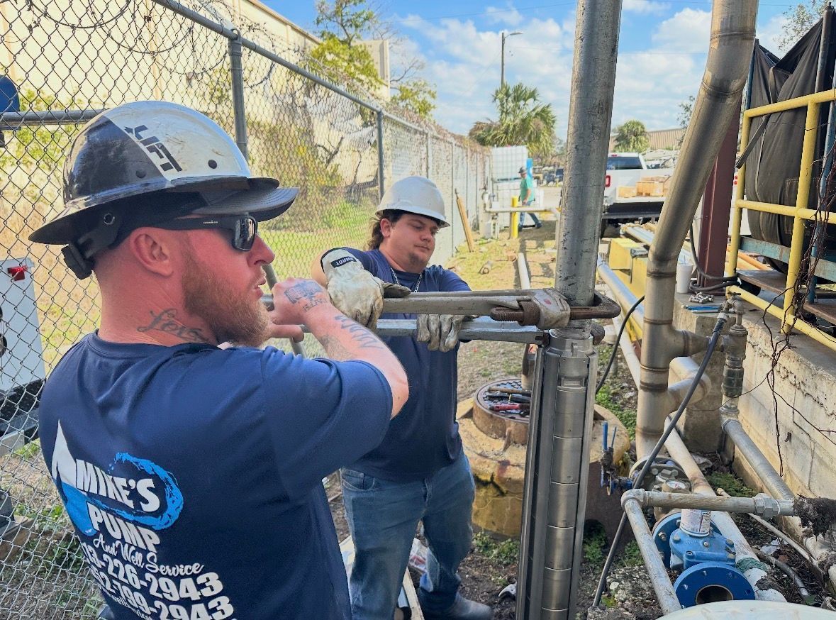 Two workers in hard hats operating a pipe valve at an industrial site near chain-link fencing.