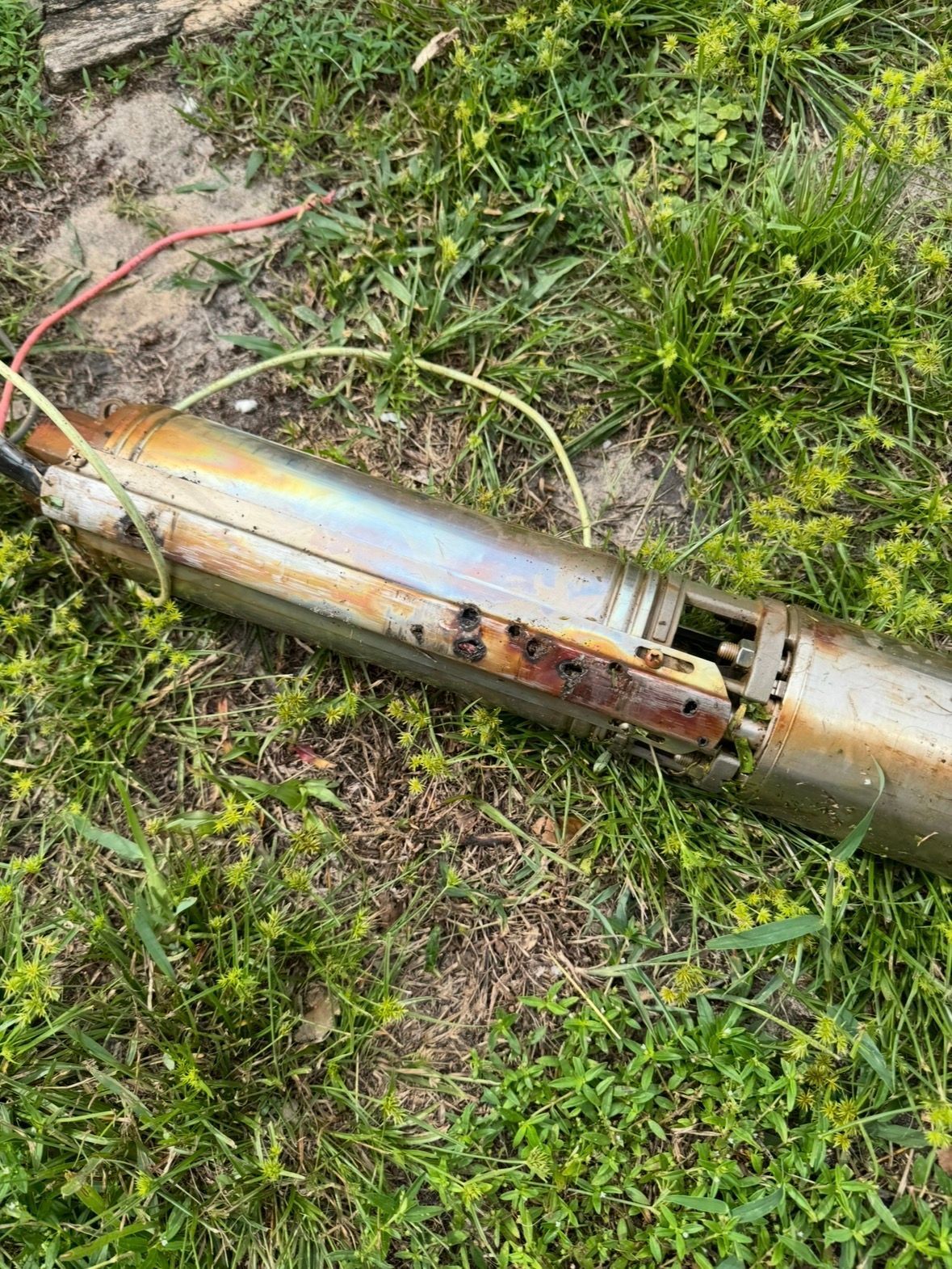 Rusty metal pipe lying in grass, with a red hose and exposed damaged section.