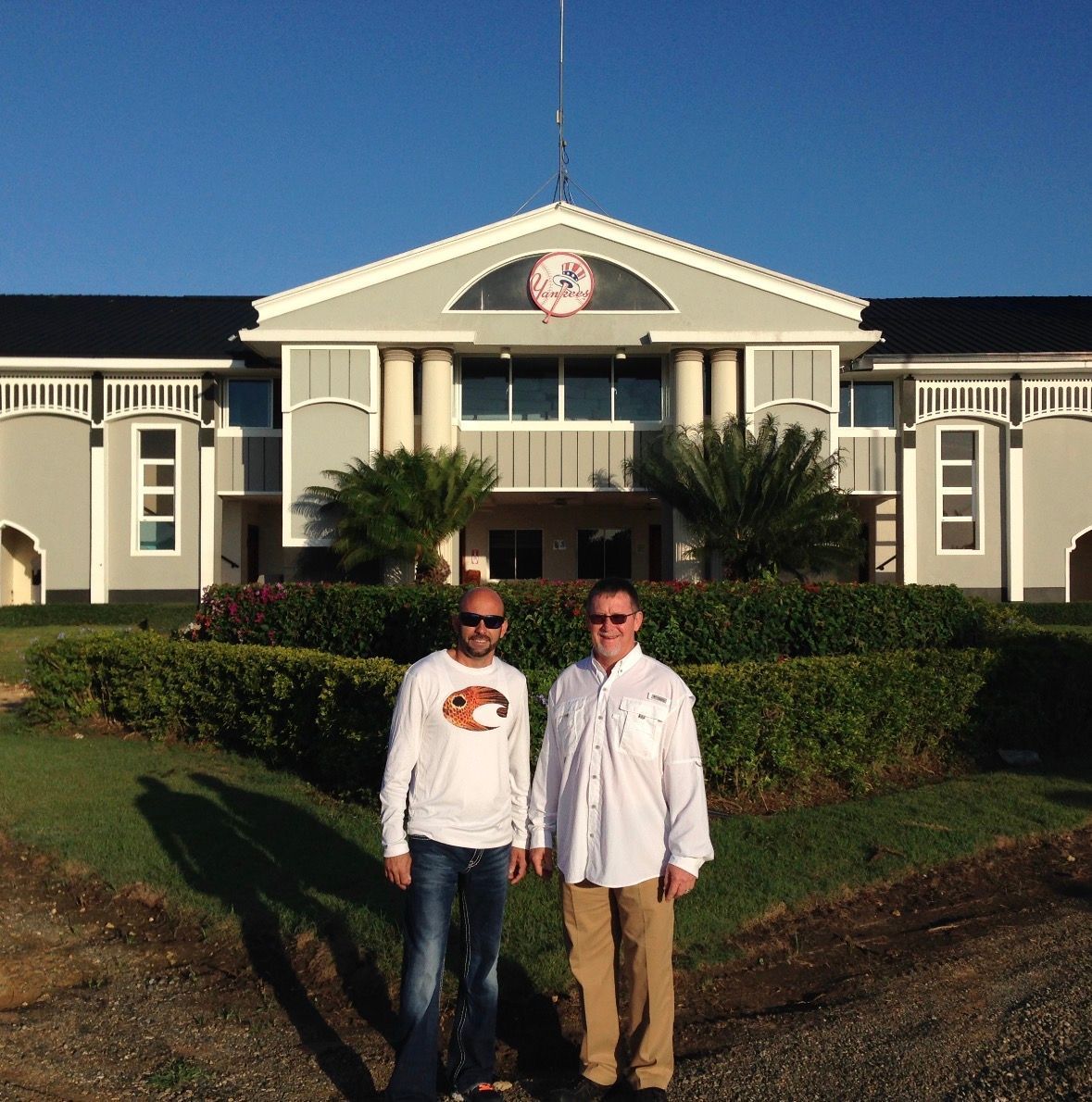 Two men standing in front of a large cream-colored building with a peaked roof on a sunny day