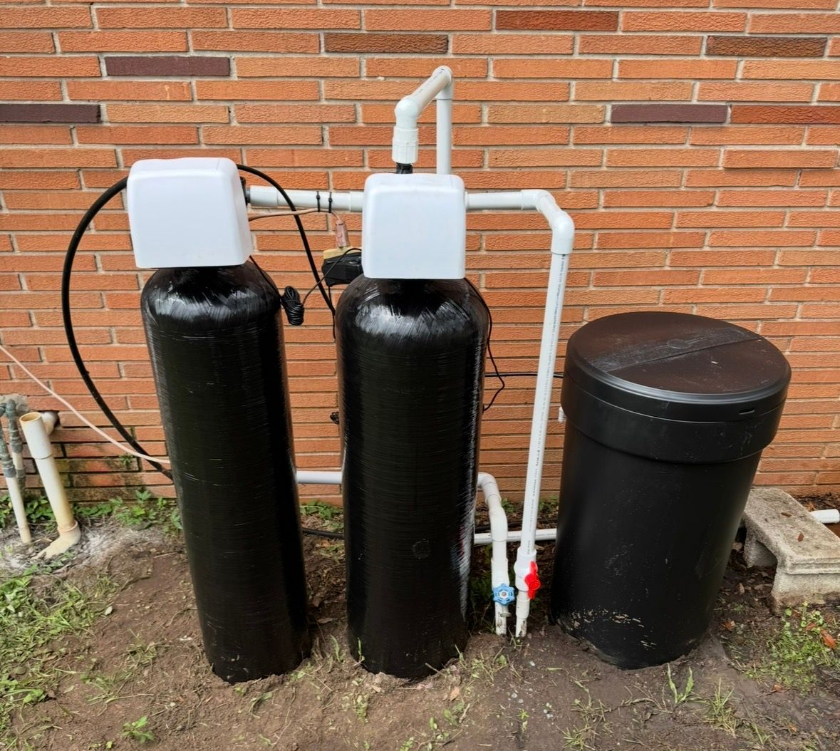 Three black water treatment tanks and a black brine tank beside a brick wall with white plumbing pipes.