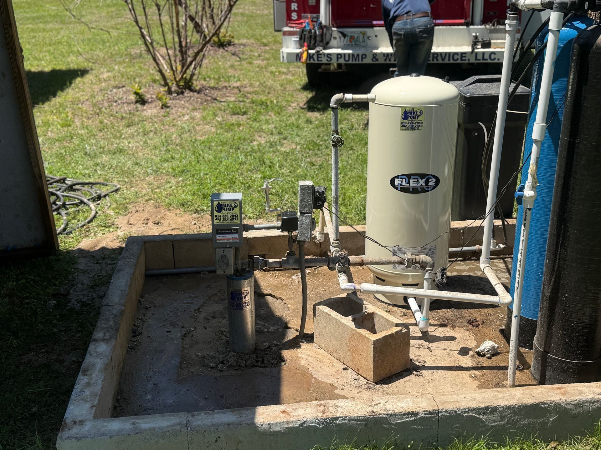 Outdoor well pump and pressure tank setup beside a house, with pipes, electrical box, and cinder blocks.