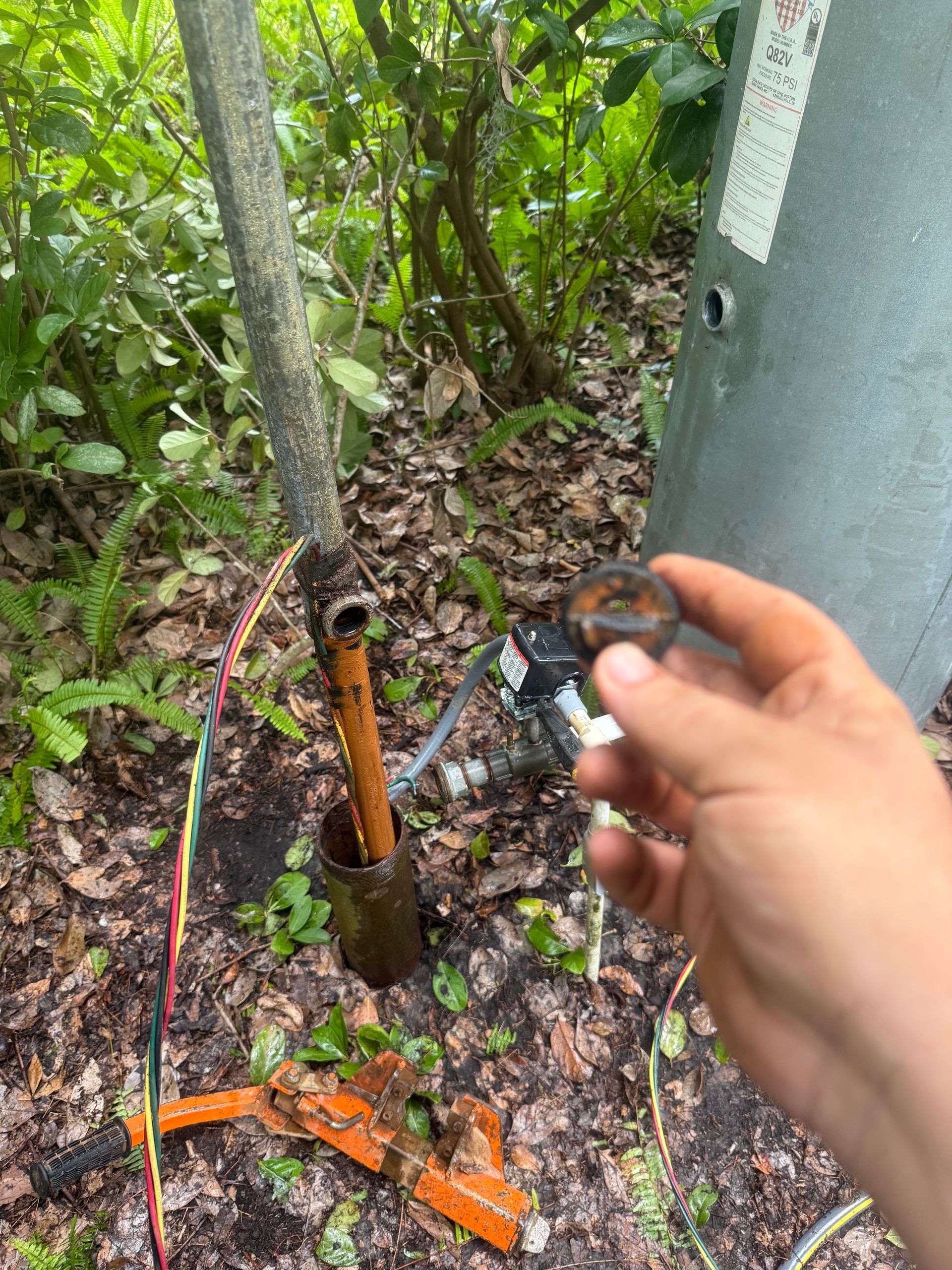 Hand holding a small pipe part beside a rusted metal post and orange debris in a wooded area