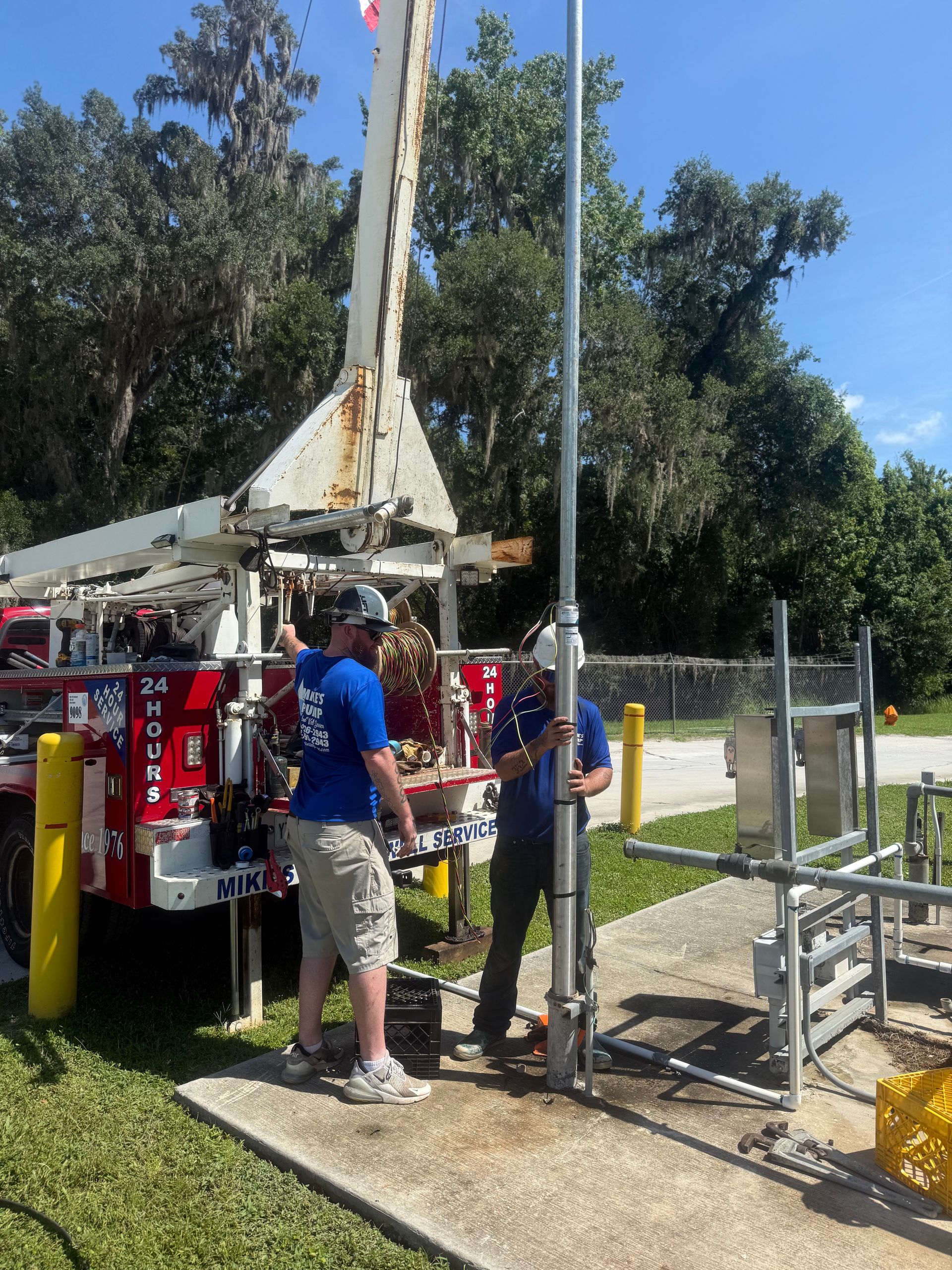 Workers beside a red drilling rig at a fenced outdoor site, with hoses and trees in the background