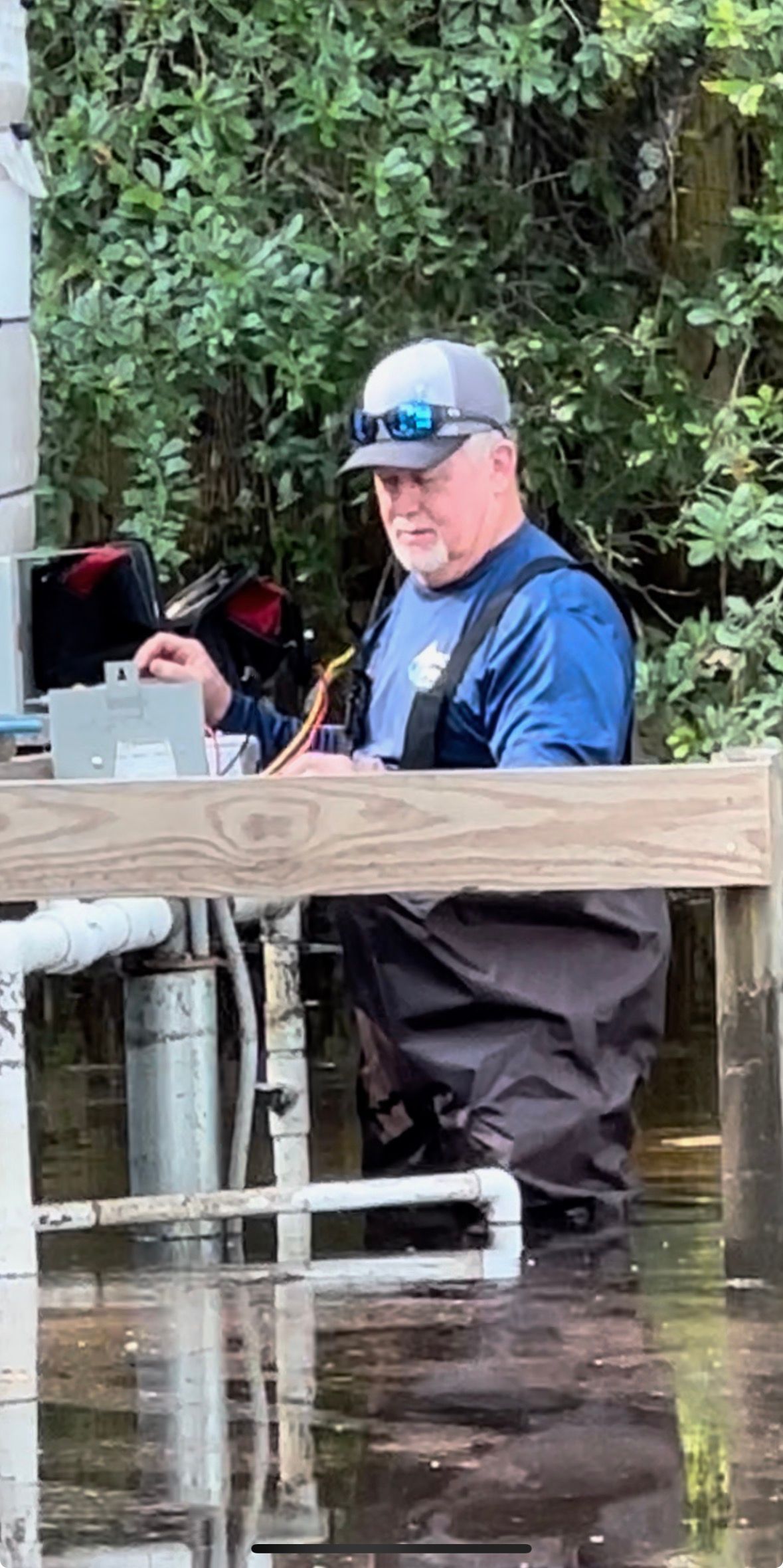 Man in blue cap and waders standing in floodwater beside a wooden barrier and equipment.