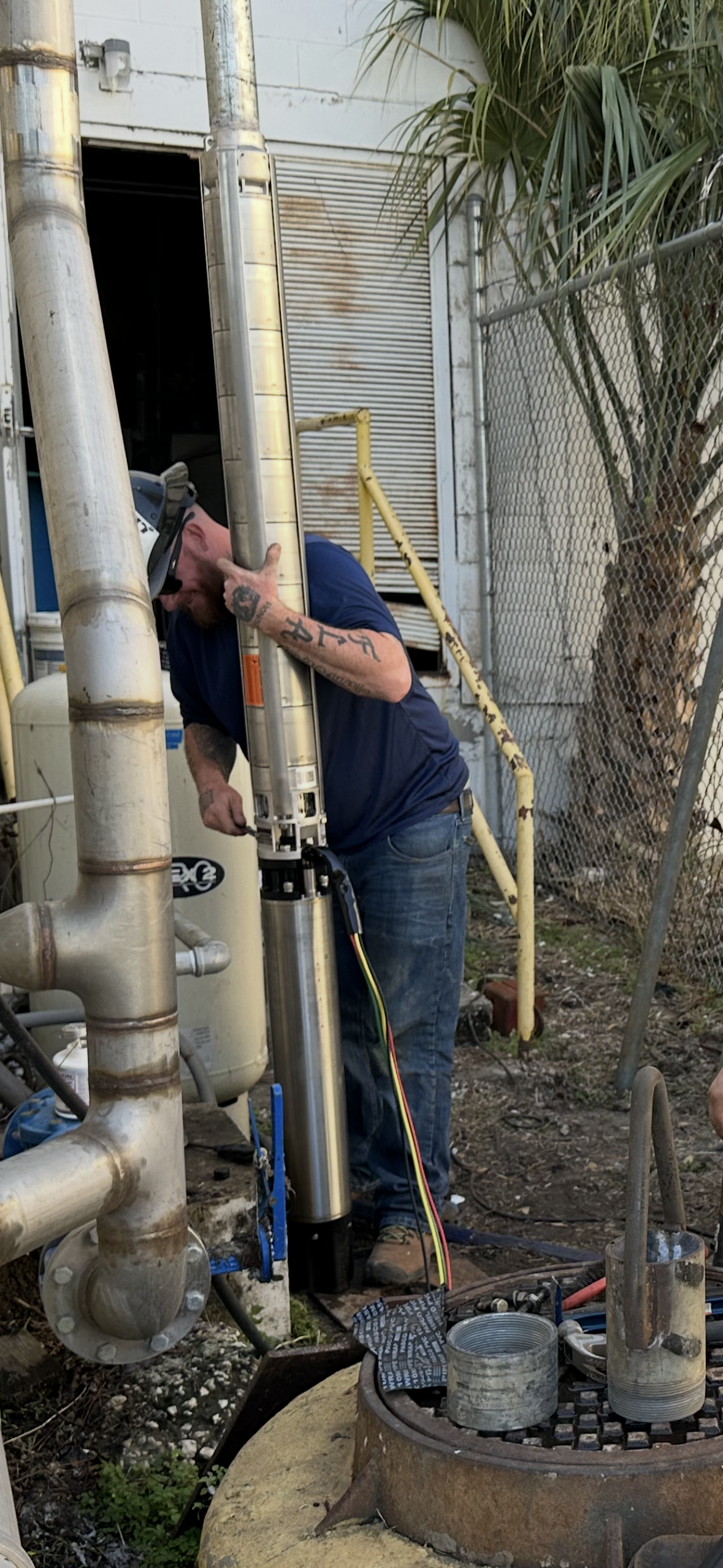 Worker inspecting pipes beside a metal building, using a tool in a narrow outdoor utility area.