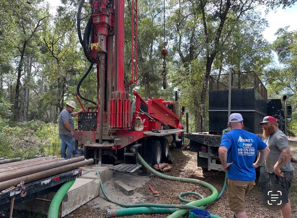 Workers operating a red drilling rig beside stacked pipes in a wooded outdoor site