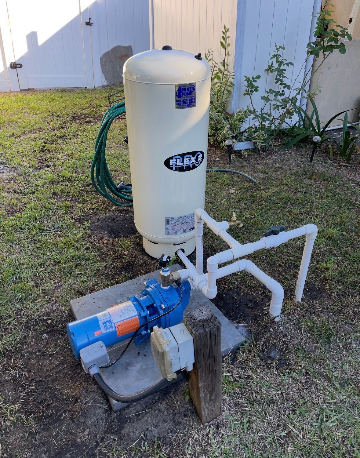 Outdoor well pump and beige pressure tank beside a building, with hoses and pipes on a small concrete pad